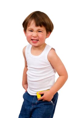 A young boy in a white baby singlet and blue jeans, smiling with a playful expression, hands in pockets.