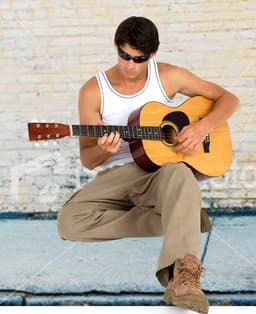A man wearing a white cotton rib singlet and sunglasses plays an acoustic guitar, sitting against a brick wall.