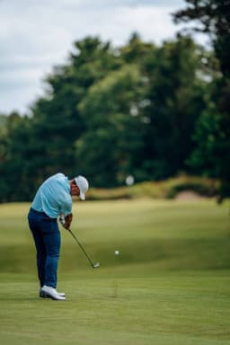 A golfer in a light blue shirt and dark trousers swings at a golf ball on a green course surrounded by trees.