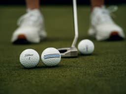 A set of three golf balls in blue and white on a green putting green, with a putter in the background.