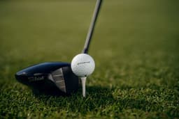 A golf club and a white golf ball on a tee, set against a green grass backdrop. The club features a logo.