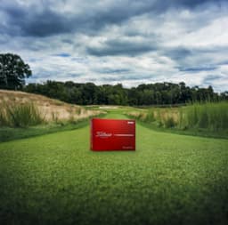 A red box containing a dozen Titleist TruFeel golf balls, positioned on a green golf fairway.