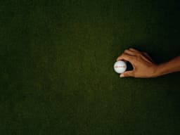 A hand holding a white golf ball on a green surface, likely artificial grass.
