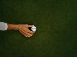 A hand holding a golf ball on a green surface, showcasing the ball’s white and coloured design.