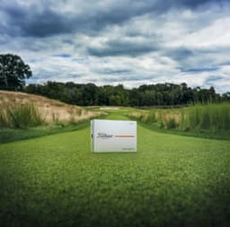 A box of Titleist 26 Velocity golf balls in white, placed on a green golf fairway under a cloudy sky.