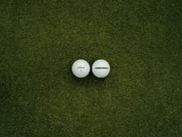 Two white golf balls are positioned on a green grass surface, featuring a logo on one ball.