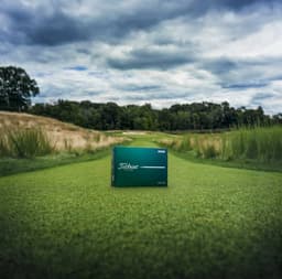 A green box containing a dozen Titleist golf balls on a grassy fairway, set against a cloudy sky.