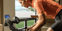 A person in an orange shirt using a bike trainer indoors, with a fan and a device displaying a training app.