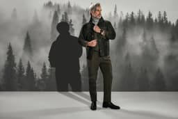 A model in a dark outfit stands against a misty forest backdrop, featuring a stylish jacket and trousers.