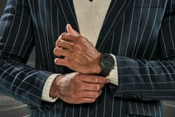 A wristwatch with a black design is displayed on a person's wrist, featuring a striped suit and cream knitted sweater.