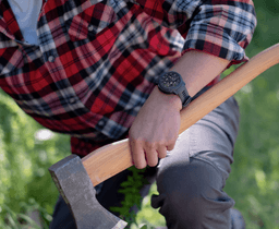A wristwatch with a black band is worn on a person's wrist, holding an axe in a green outdoor setting.