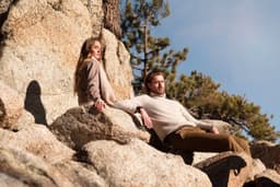 A man and a woman sit on large rocks outside, surrounded by trees and a blue sky. The woman wears a light sweater, and the man is in a light-coloured knit.