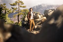 A person stands among boulders in a natural landscape, wearing a brown coat and tan pants. Trees and mist surround them.