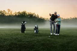 A golfer instructs a young player on a misty field at sunrise, with golf bags in the background.