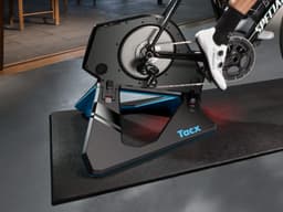 A smart trainer in black and blue with a visible bike mounted on it, set on a mat in an indoor space.