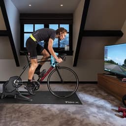 A cyclist using a black Garmin Tacx FLUX S Smart Trainer in a well-lit indoor space, facing a TV screen.