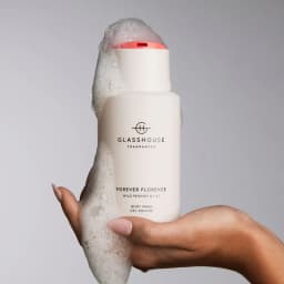 A white bottle of body wash with a red cap, featuring bubbles, held in a hand. It has a logo and branding.