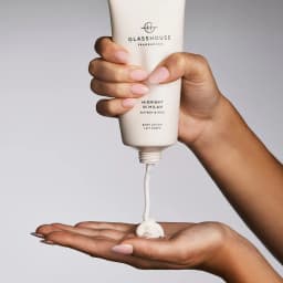 A white tube of Glasshouse Body Lotion Midnight in Milan is being squeezed, dispensing a creamy moisturiser into a hand.