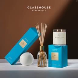 A diffuser and a candle in blue and gold packaging, accompanied by a white sphere, on a neutral surface.