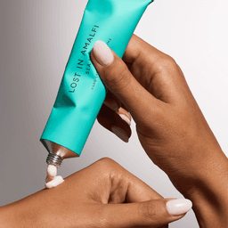 A hand applying white cream from a turquoise tube labelled "Lost In Amalfi". The hand has neatly manicured nails.