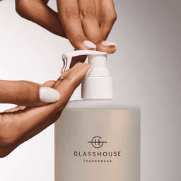 A hand is dispensing a clear liquid from a glass hand wash bottle with a white pump and a simple logo.