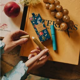 A person holds two perfume pencils, one gold and one turquoise, alongside a floral-patterned box on a textured yellow surface.