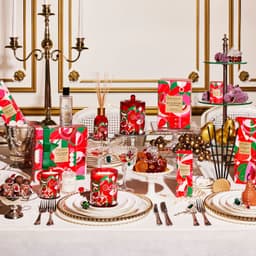 A festive table setting featuring various candles and fragrance products in red and green packaging, displayed elegantly.