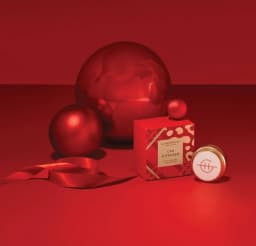 A red car diffuser with packaging, surrounded by red ornaments and ribbon, set against a solid red background.