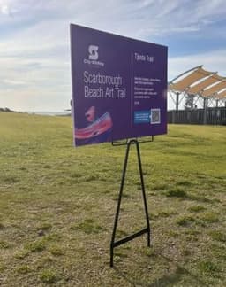 A promotional display featuring a purple sign with information about the Scarborough Beach Art Trail, on a black wire stand.