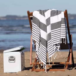 A grey and white striped Patara Turkish towel draped over a wooden beach chair beside a cooler on the sand.