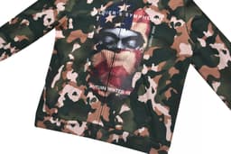 A camouflage-patterned hoodie featuring a direct print graphic of a masked figure against an American flag backdrop.