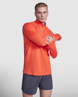 An orange half zip sweatshirt is worn by a model with short hair, paired with dark shorts. The garment includes a logo.