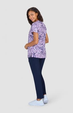 A woman wearing a purple patterned top and navy trousers stands facing sideways, smiling at the camera.