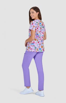 A woman wearing a floral print top in pink and purple, paired with lilac pants and white shoes, standing against a grey background.