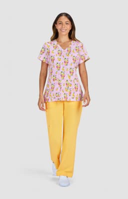 A woman wears a pink patterned top featuring floral designs and yellow trousers, posing against a grey background.
