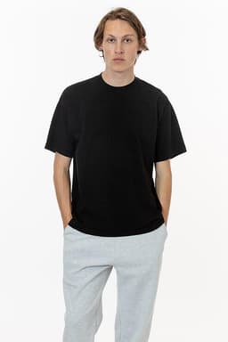A black short sleeve crewneck t-shirt worn by a model, paired with light grey pants. The t-shirt features a logo.