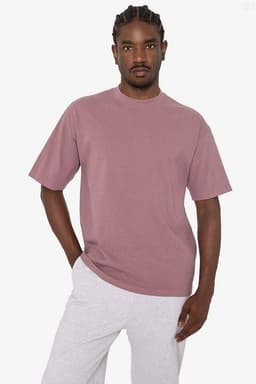 A man wearing a short sleeve crewneck tee in a muted mauve colour, paired with grey pants. The tee features a logo.