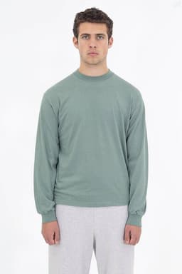 A long sleeve crewneck tee in a soft green hue, worn with light grey bottoms. The shirt features a logo on it.