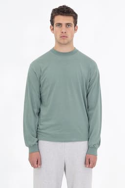 A long sleeve crewneck tee in a soft green colour, featured on a model wearing grey pants. The shirt has a logo.