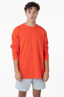 A model wearing an orange long sleeve crewneck tee paired with grey shorts, standing against a plain background.