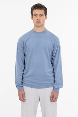 A long sleeve crewneck tee in a soft blue colour, worn by a model with grey pants. It features a small logo.