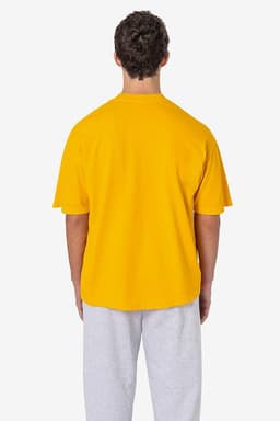 A man stands with his back to the camera, wearing a bright yellow short sleeve pocket tee and grey sweatpants.