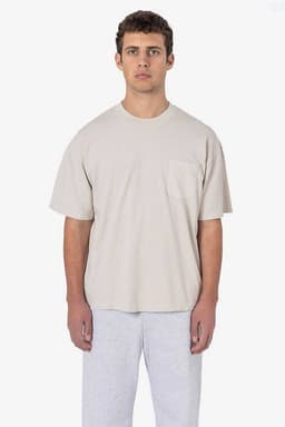 A model wears a beige short sleeve garment-dyed pocket tee paired with grey trousers. The shirt features a pocket.