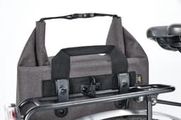 A grey cycle bike bag with a handle and fastening, attached to a bike rack. It has a sturdy design and a logo.