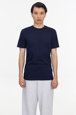 A man stands wearing a navy short sleeve fine jersey crew tee and grey sweatpants. The shirt features a logo.