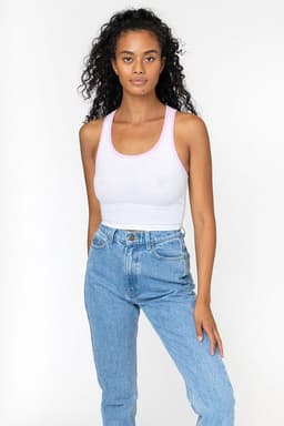 A woman models a white rib crop tank top with pink accents, paired with high-waisted blue jeans. The top features a logo.