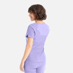 A woman in a lilac Y-neck top is smiling, showcasing the back view of the shirt. The top has a logo.