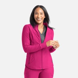 A woman wearing a bright pink full zip track jacket, smiling and standing against a light grey background.