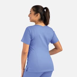 A woman stands with her back to the camera, wearing a light blue double V-neck top. The top features short sleeves and has a logo.
