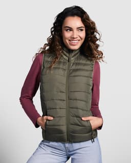 A woman models an olive green padded vest, featuring a zip front and side pockets, paired with a burgundy long-sleeve top.
